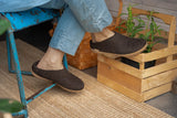 Person wearing brown slippers on a woven floor with a wooden crate and plant in the background.