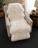 A fluffy white sheepskin-style throw blanket draped over a light beige armchair in a softly lit room with a side table, lamp, plant, and dark carpet visible.